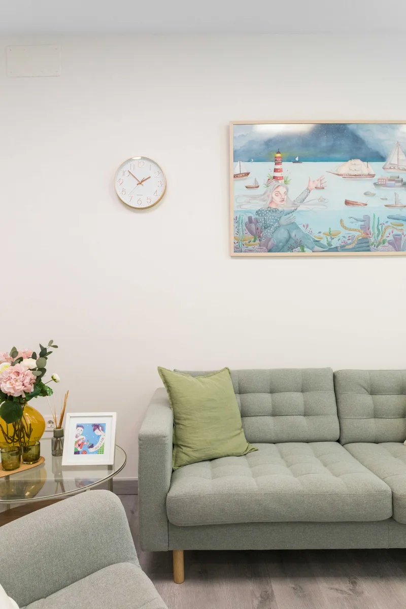 Detalle del salón: sofá verde con cojines y flores frescas bajo una lámina marina